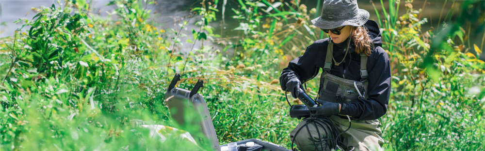 a person in overalls kneels among tall lgrasses and removes a monitoring device from a large case