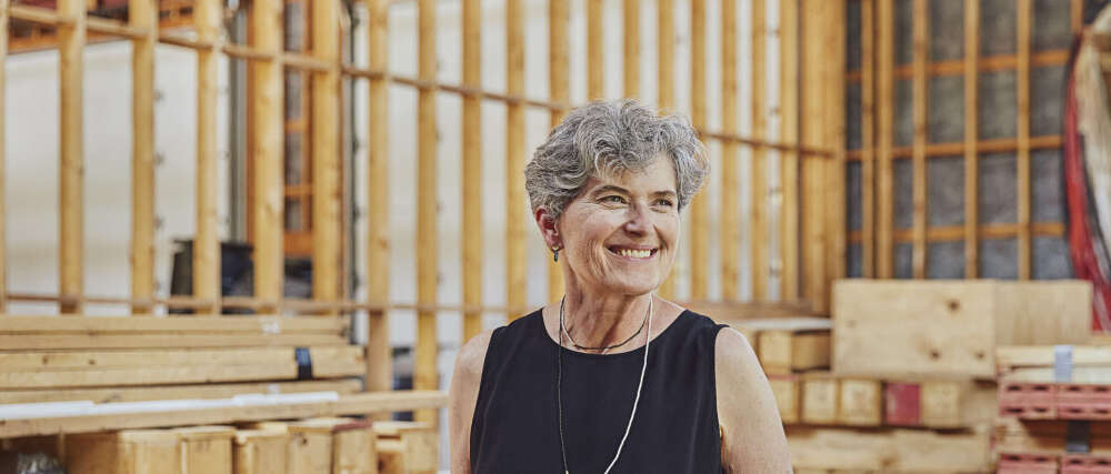 A person with short grey hair wearing a black sleeveless shirt and a silver necklace stands smiling facing left in a workspace with wood behind them.