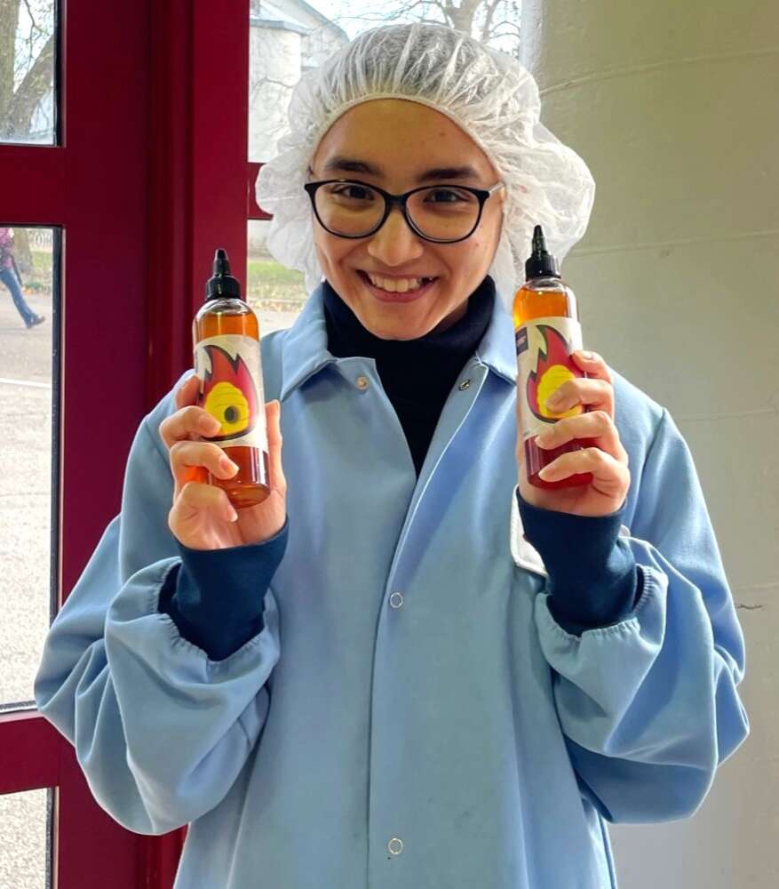 a person in a lab coat and hair net smiles while holding two bottles of honey