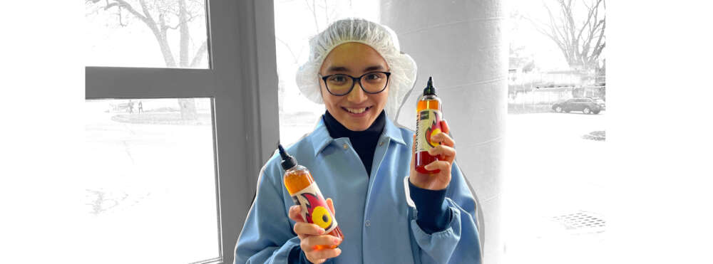 a person in a lab coat and hair net smiles while holding two bottles of honey