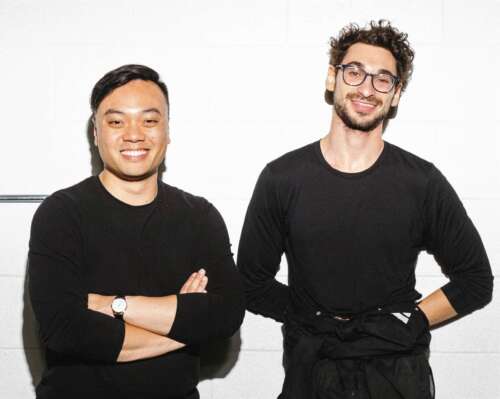 Two men wearing all black stand beside one another smiling into the camera in front of a white background.