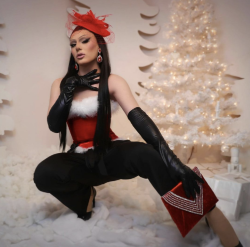 A person dressed in Christmas-themed drag hair, makeup and clothing crouches on a white rug holding a red purse in front of a lit up white Christmas tree.