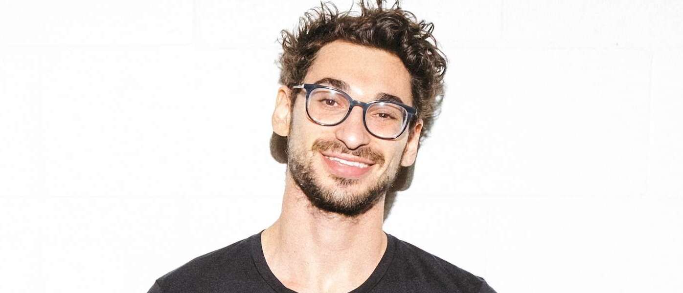 A person with curly black hair, black eyeglasses and black facial hair wearing a black t-shirt smiles standing in front of a white background.