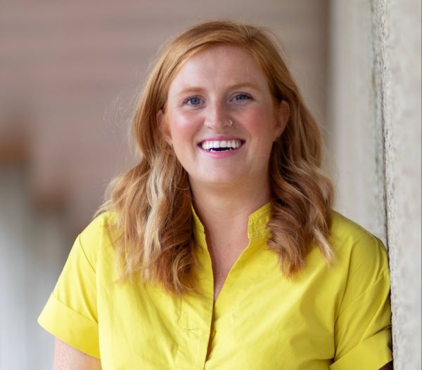 a person smiles while posing for a photo while leaning against a concrete building