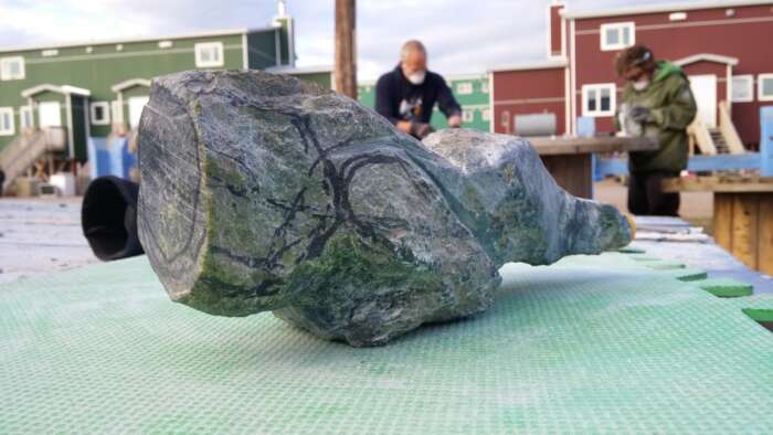 A grey object with hints of green and yellow with black lines drawn over it. The surface of the object appears to be rough and hard. It rests on a piece of green foam. In the background, a green and red house are visible along with two people who appear to be working.