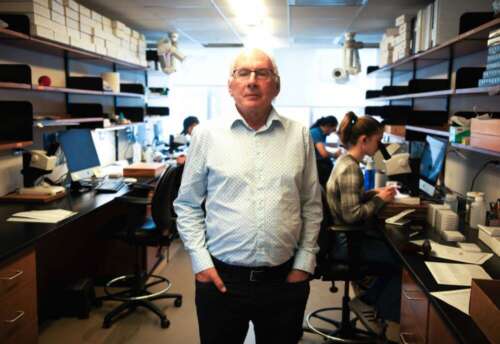 A man stands in a lab as workers behind him peer into microscopes