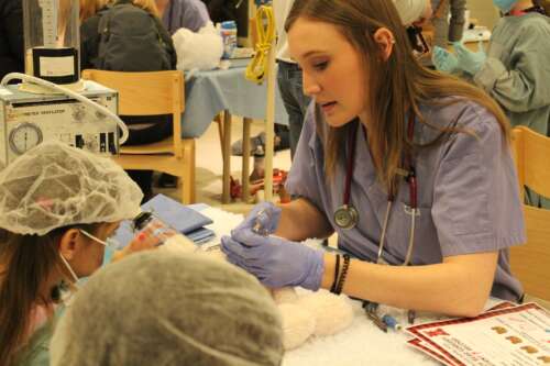 A student dressed as a vet repairs a stuffed animal in front of two children during College Royal.