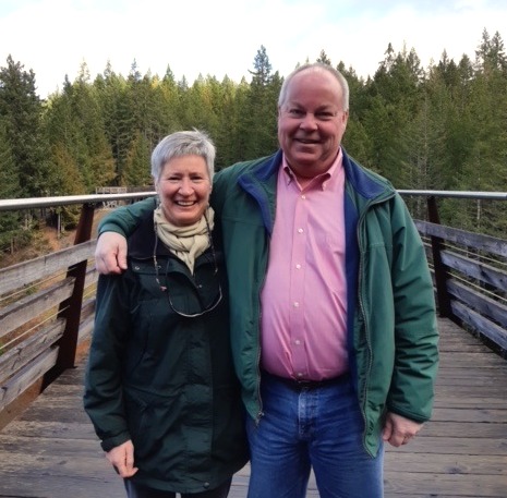 A man has his arm around a woman as they stand on a boardwalk with a forest behind