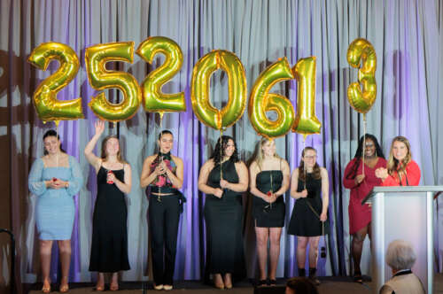 A woman at a podium while 7 women in formal wear hold balloons with the numbers 2520613