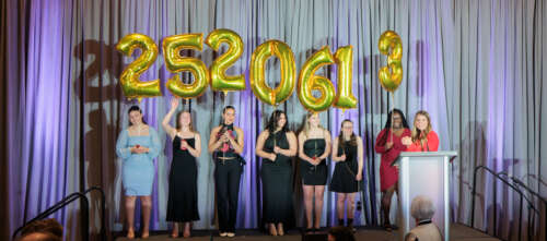 Seven women in formal attire stand on a podium holding balloons with the number 2520613