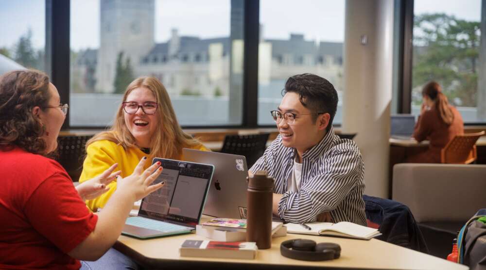 Two people sitting at a desk with laptops before them laugh while a third person at the desk gesticulates while speaking