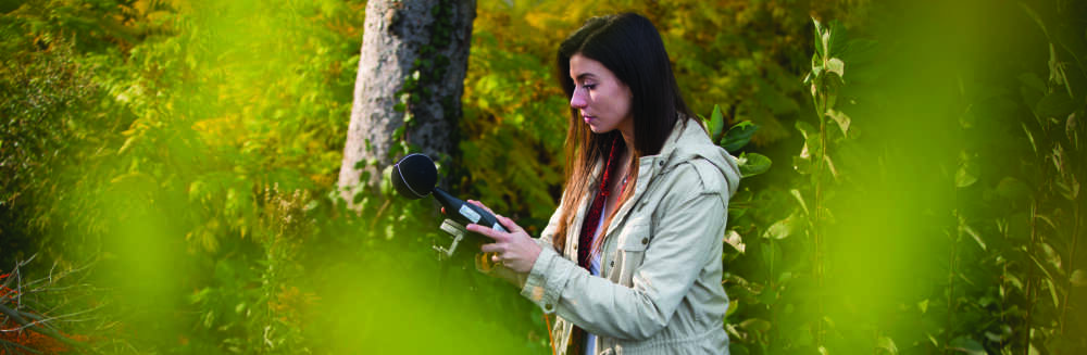 A woman looks at a surveying device while standing in the woods.