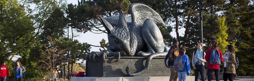 A group of students chat while standing by the Gryphon statue on a sunny day.