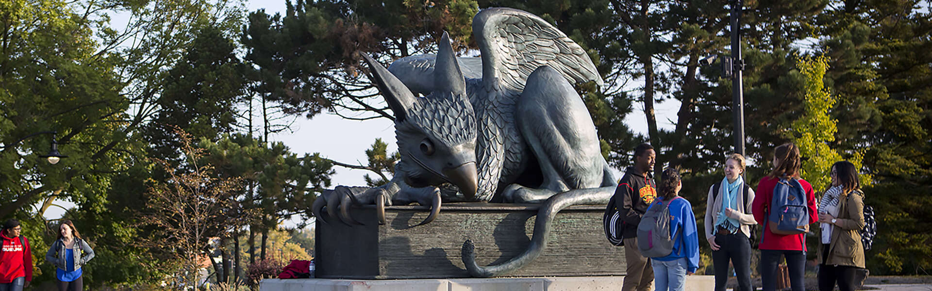 A group of students chat while standing by the Gryphon statue on a sunny day.