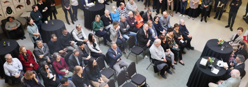 View from second floor of the recognition event in the Centre for Biodiversity Genomics. Roughly 50 attendees sit in chairs in the middle while another 50 stand around the perimeter. Dr. Paul Hebert and panel sit at round high tables at the front of the room.