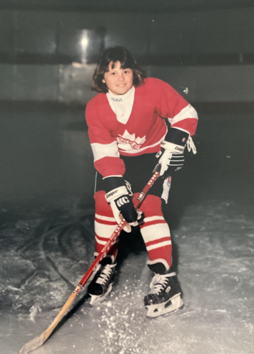 A young girl with brown hair and bangs stands on the ice in position, dressed in red, white and black hockey gear, holding her stick and smiling at the camera.