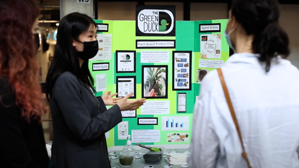 Student presenting a poster display labelled as "The Green Duck" and various sample products on table