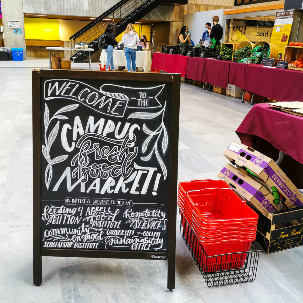 A sign advertises the campus food market in the University Centre Courtyard. Set up in the courtyard are tables filled with produce for sale.