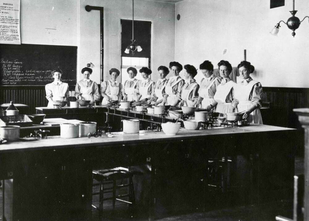 Eleven people in OAC's cooking classroom in 1915, dressed in aprons and standing around a cooking workstation