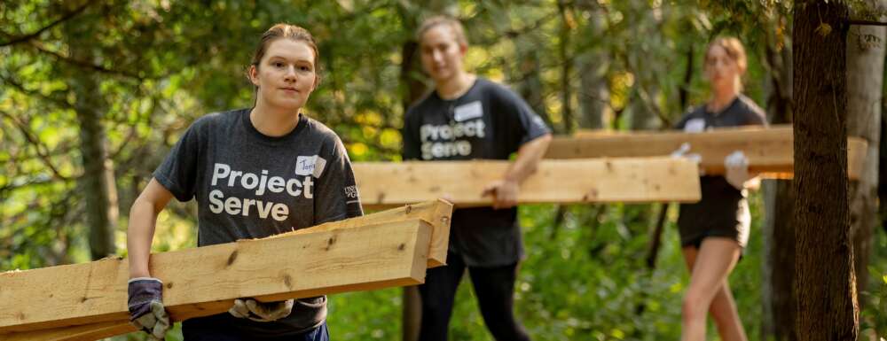 Students wearing project serve tshirts carry long wooden planks through a wooded trail.