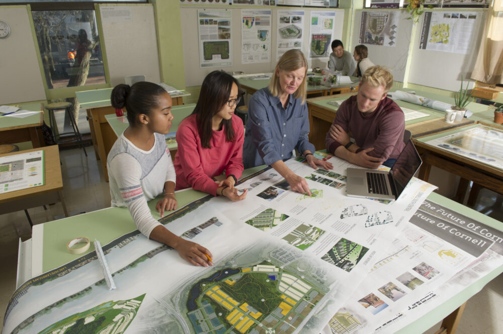 Four people standing around a map in OAC's landscape architecture classroom; a teacher points at a diagram as students study intently