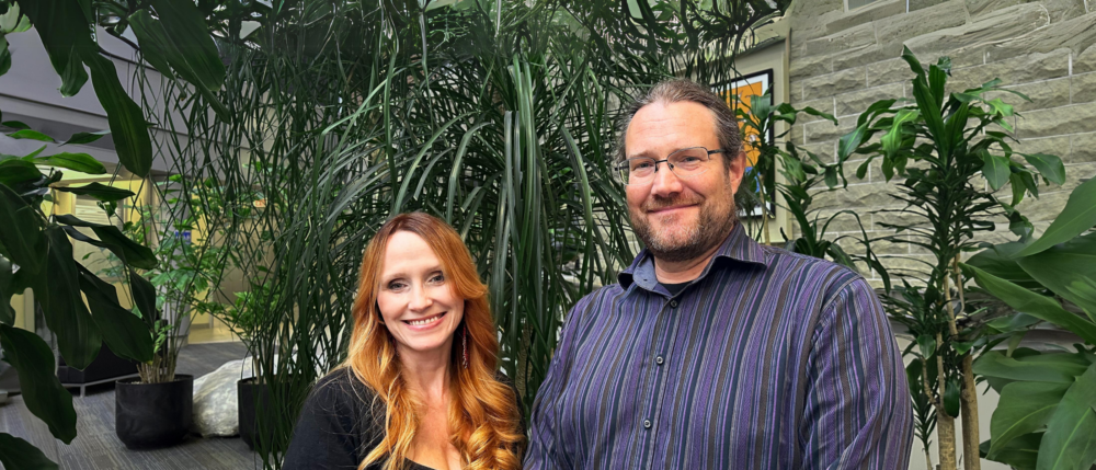 A person wearing black with long red hair stands in front of a lush green plant with a person with a beard in a blue striped shirt wearing glasses.