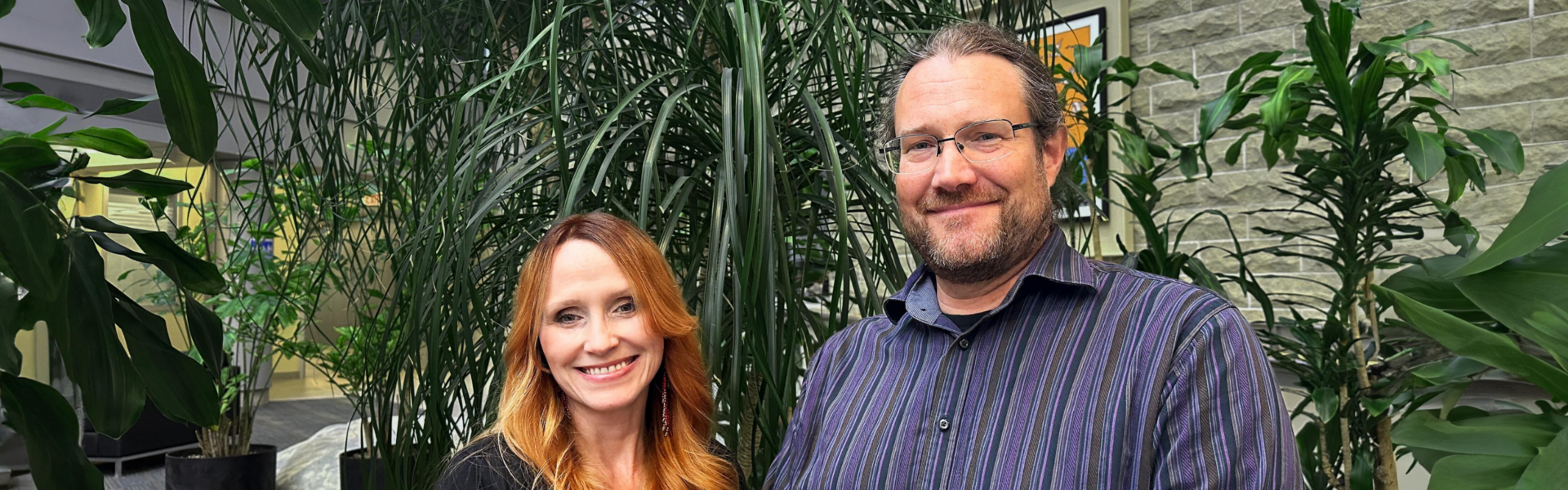A person wearing black with long red hair stands in front of a lush green plant with a person with a beard in a blue striped shirt wearing glasses.
