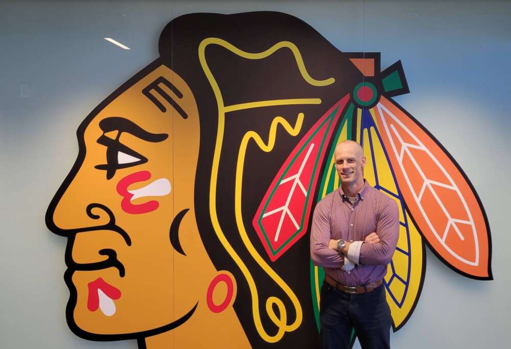 A ma stands smiling in dark pants and a red plaid button-up shirt with his arms crossed, in front of a wall that depicts the Chicago Blackhawks logo.