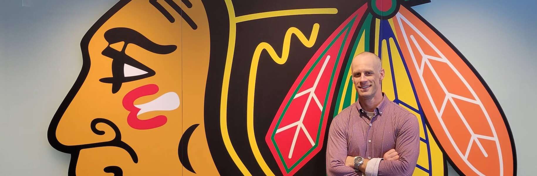 A ma stands smiling in dark pants and a red plaid button-up shirt with his arms crossed, in front of a wall that depicts the Chicago Blackhawks logo.