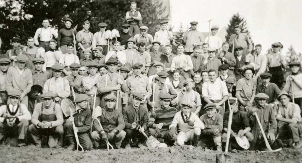 Black and white photo of several dozen labourers holding tools pose for a group shot,