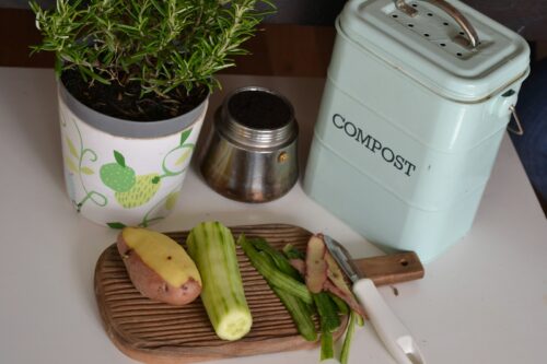 A cutting board with vegetables and knife and a metal compost bin behind