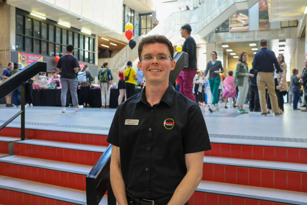 Adam Morris stands at the base of a set of steps in the University Centre and smiles at the camera wearing a black U of G polo shirt.