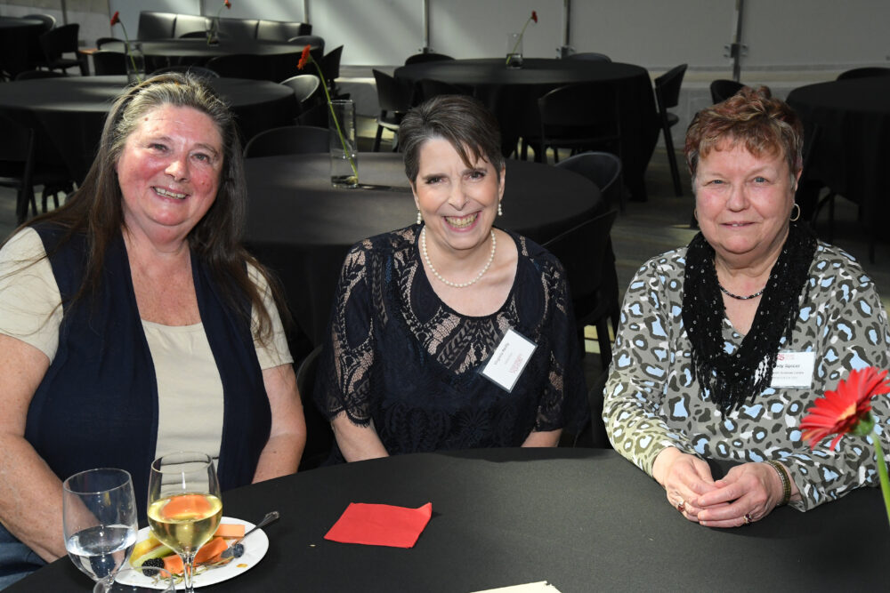 3 women sit at a table and smile at the camera.