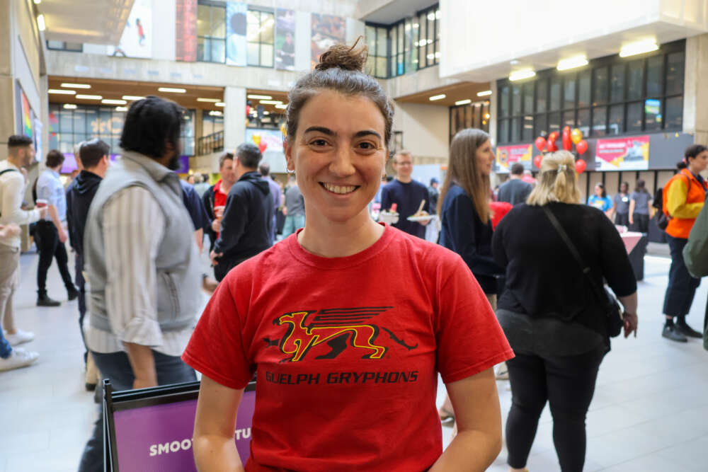 Genna Villella stands in the UC during the staff social. She's wearing a red Guelph Gryphons t-shirt and smiling at the camera.