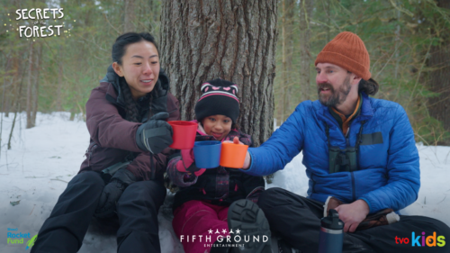 Three people in winter wear sit on the snow in a forest holding mugs of hot drinks