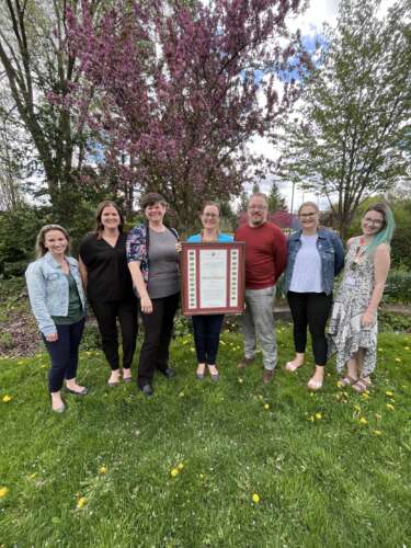 7 members of the student engagement and leadership team pose with Shannon as she holds a large framed award outside.