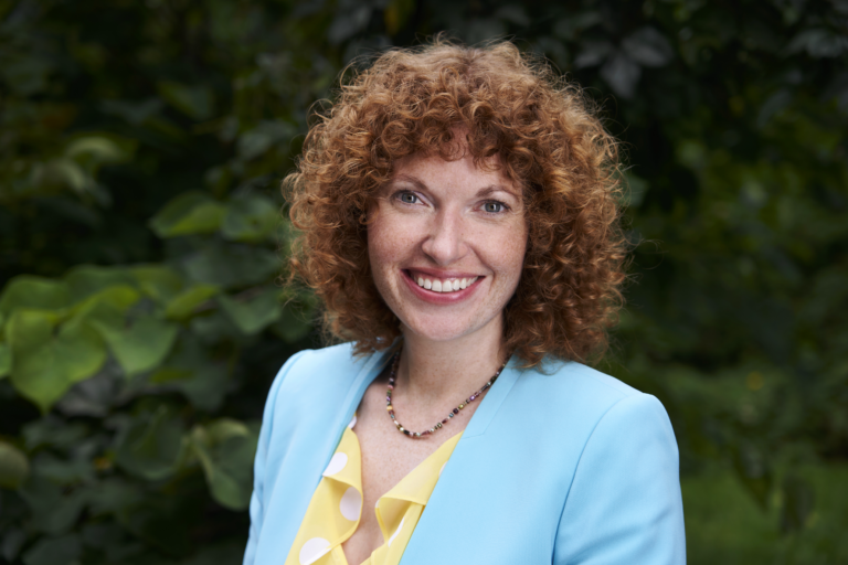 Headshot of Dr. Jennifer Geddes-McAlister against a background of plants. She has curly red hair and is dressed in a blue blazer. 