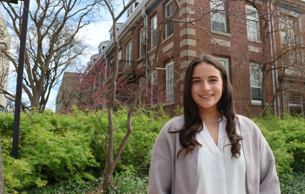 A student with long brown hair, wearing a white blouse and a grey cardigan stands smiling in front of a red brick building and landscaped greenery on campus.