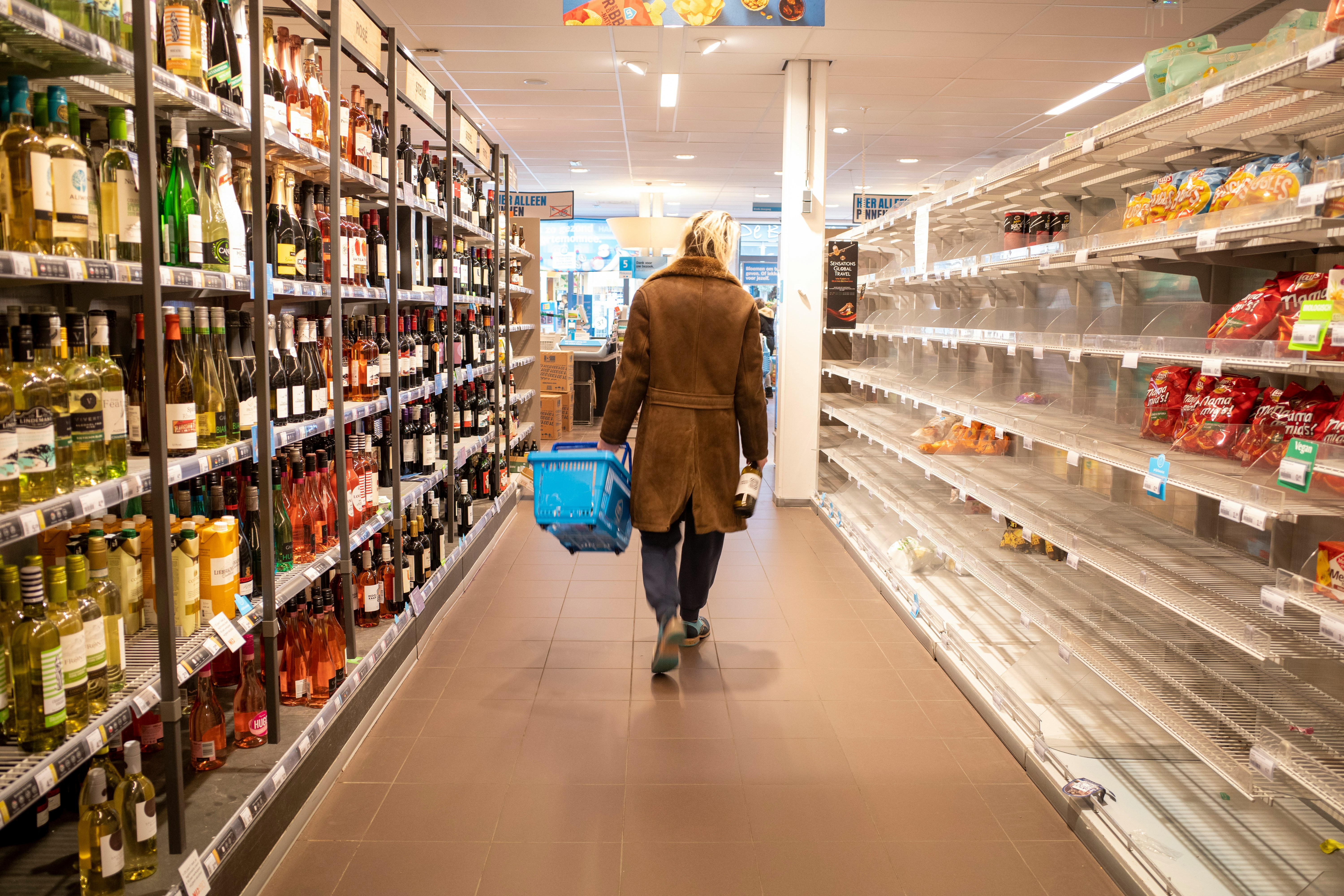 Person in browncoat walking down an aisle of a grocery store, the shelf on their left is significantly emptied of goods