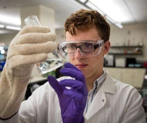 A student wearing protective lab wear examines a liquid while in a lab