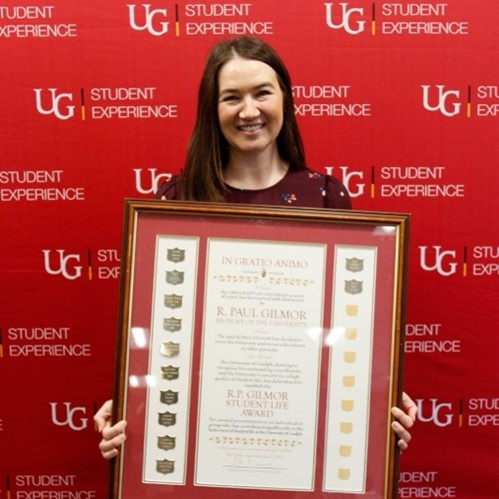 Ally holding her large framed award while standing in front of a red U of G Student Experience backdrop and smiling.