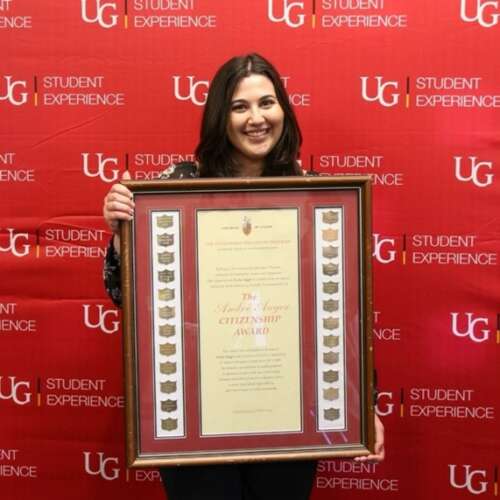 Angelica holding her large framed award while standing in front of a red U of G Student Experience backdrop and smiling. 