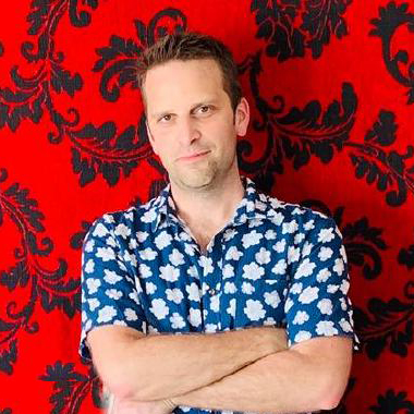 Dr. Ben Devries stands with his arms crossed in front of a bright red background. He's wearing a short sleeve blue button down shirt with a pattern of white clouds.