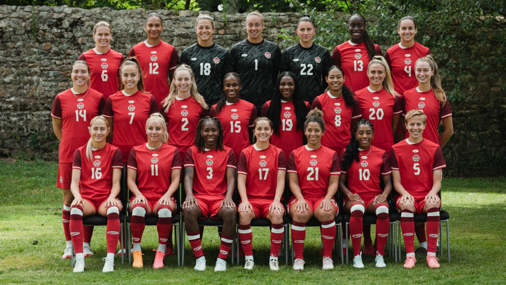 Group photo of Canada Olympic Women's Football team. People in red uniforms smile as they sit and stand in rows on a green field. 