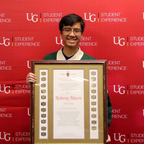 Issac holding his large framed award while standing in front of a red U of G Student Experience backdrop and smiling.