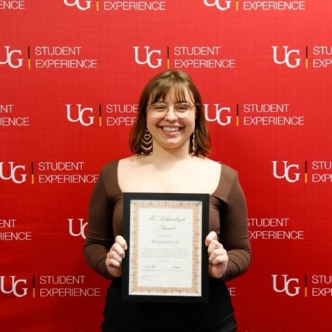 Maegan Ellis holding her large framed award while standing in front of a red U of G Student Experience backdrop and smiling.