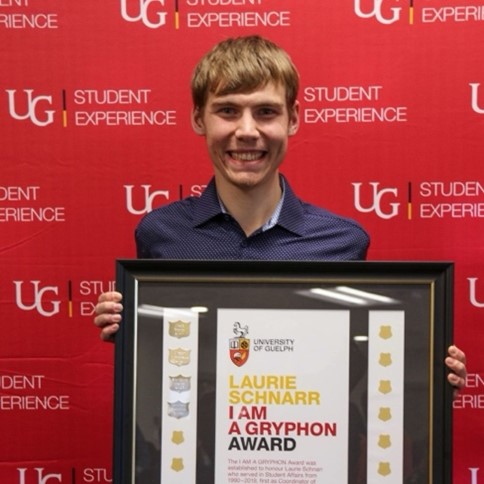 Michael Hamilton holding his large framed award while standing in front of a red U of G Student Experience backdrop and smiling.