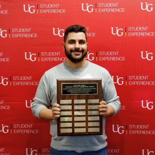 Nabhan holding his large framed award while standing in front of a red U of G Student Experience backdrop and smiling.