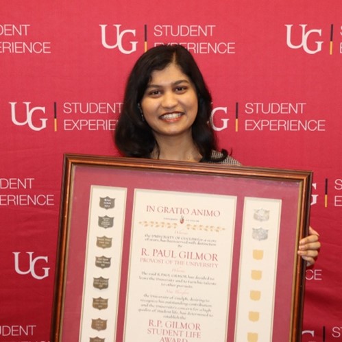 Riya holding her large framed award while standing in front of a red U of G Student Experience backdrop and smiling.