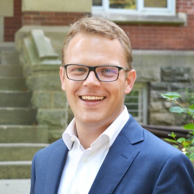 Rogier Holtermans stands in front of a red brick building dressed in a blazer and white button down shirt, smiling at the camera.